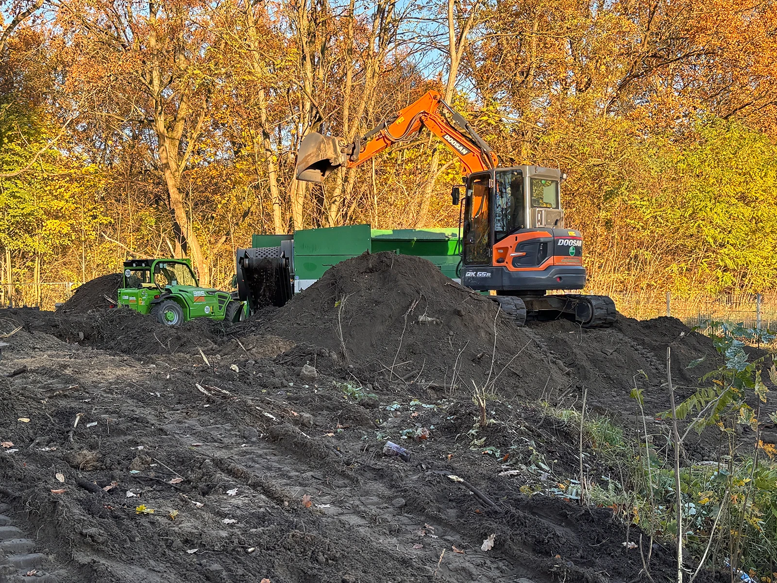 Gartenfit Wotschke UG - Bagger auf einem Erdhaufen, schaufelt Erde in eine Zemmler MS 1600 Doppeltrommel Siebanlage. Im Hintergrund ein Radlader.